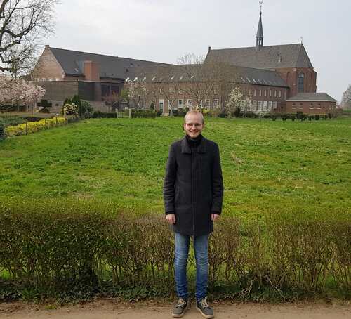 Stef Coenen staat in de kloostertuin, met op de achtergrond het klooster met Erfgoedcentrum Nederlands Kloosterleven. Bron Erfgoed Brabant.