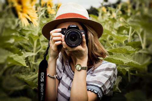 Jonge vrouw tussen de zonnebloemen maakt een foto richting de kijker. Foto Andre Furtado. Bron Pexels.
